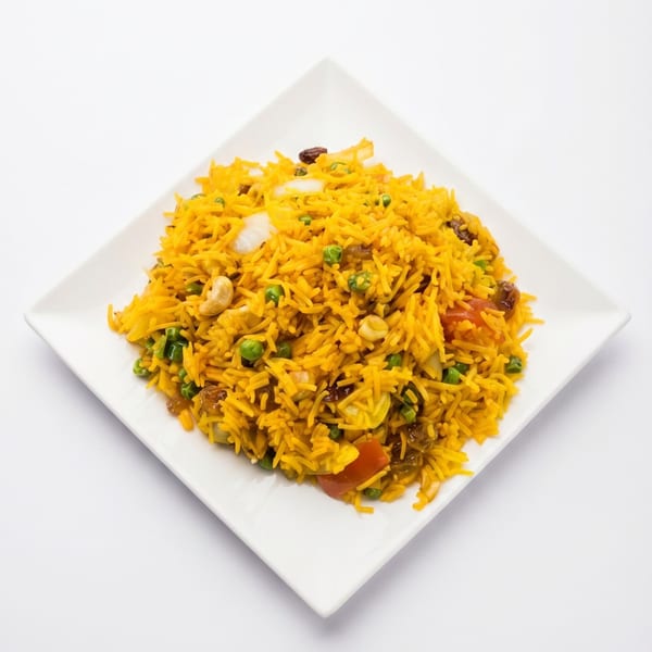 A square white plate filled with yellow rice, green peas, cashews, raisins, and red bell pepper.