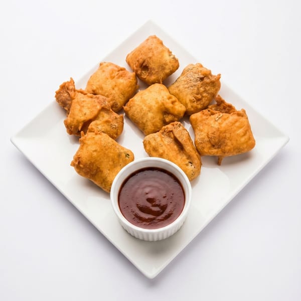 A white square plate holds eight golden-brown fried pakoras and a small bowl of dark red chutney.